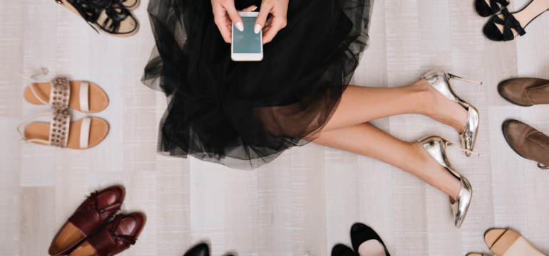 Stylish girl sitting on the floor in a dressing room with smartphone in hands, writes the message, surrounded by a variety of shoes. She is dressed in a black skirt, on her feet silver luxury shoes.